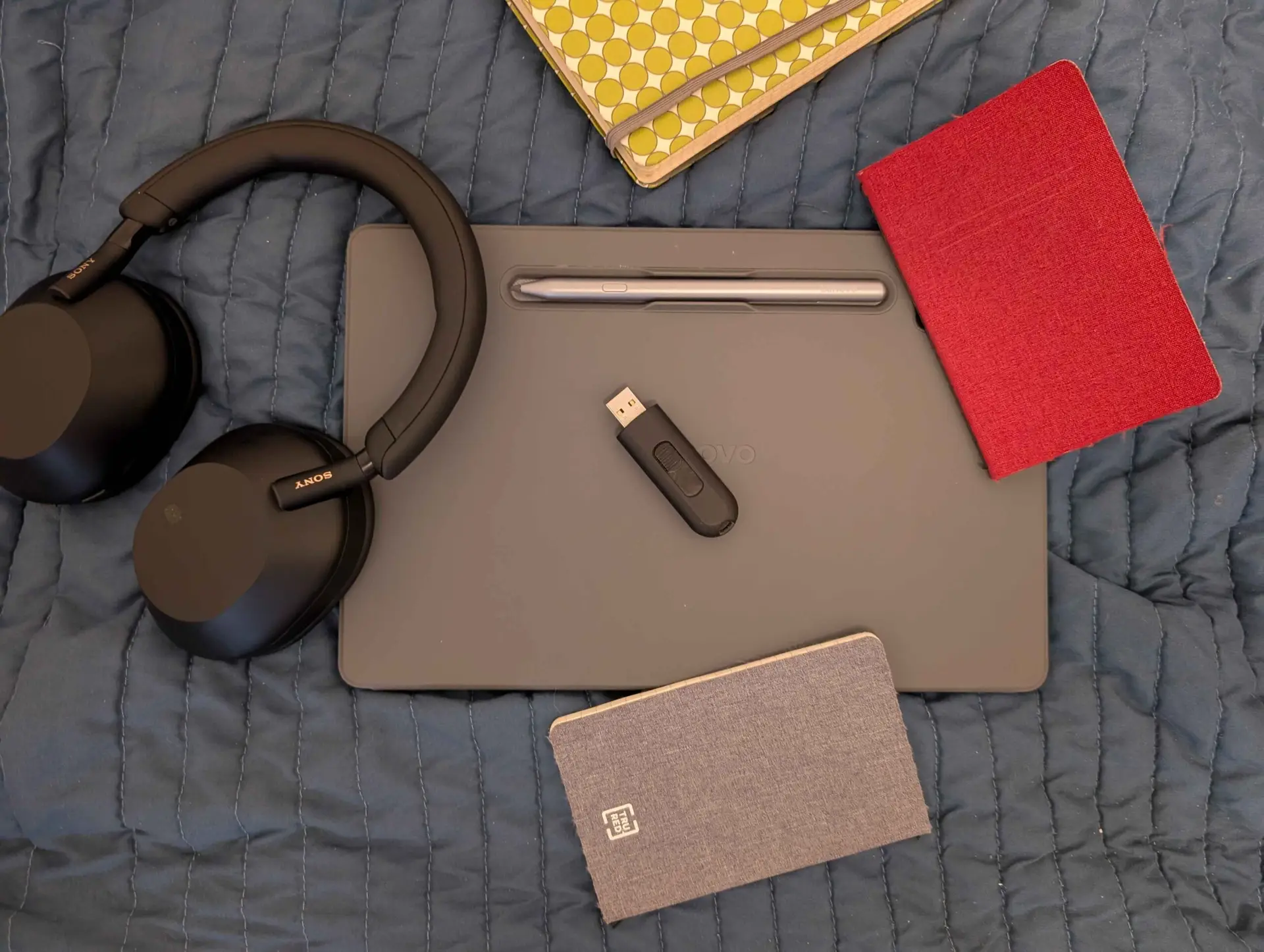 A USB flash drive sits on top of a Lenovo tablet, surrounded by notebooks and Sony headphones.