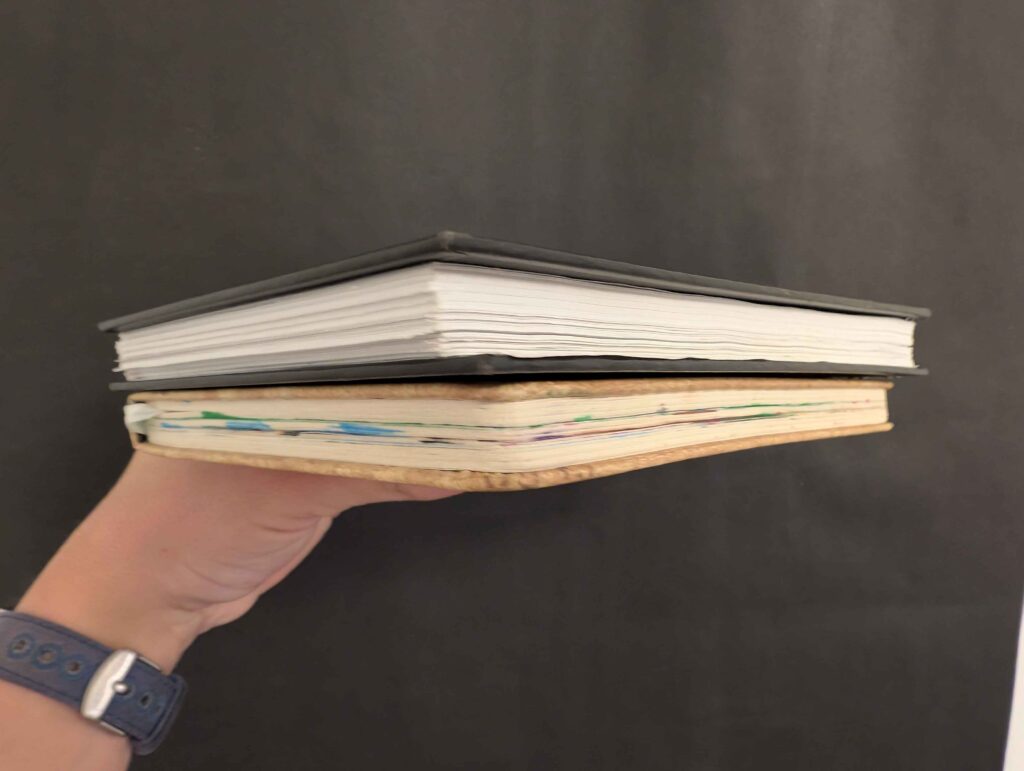 A man's hand holding up two art notebooks, one with clean page edges and the other colorful pages.