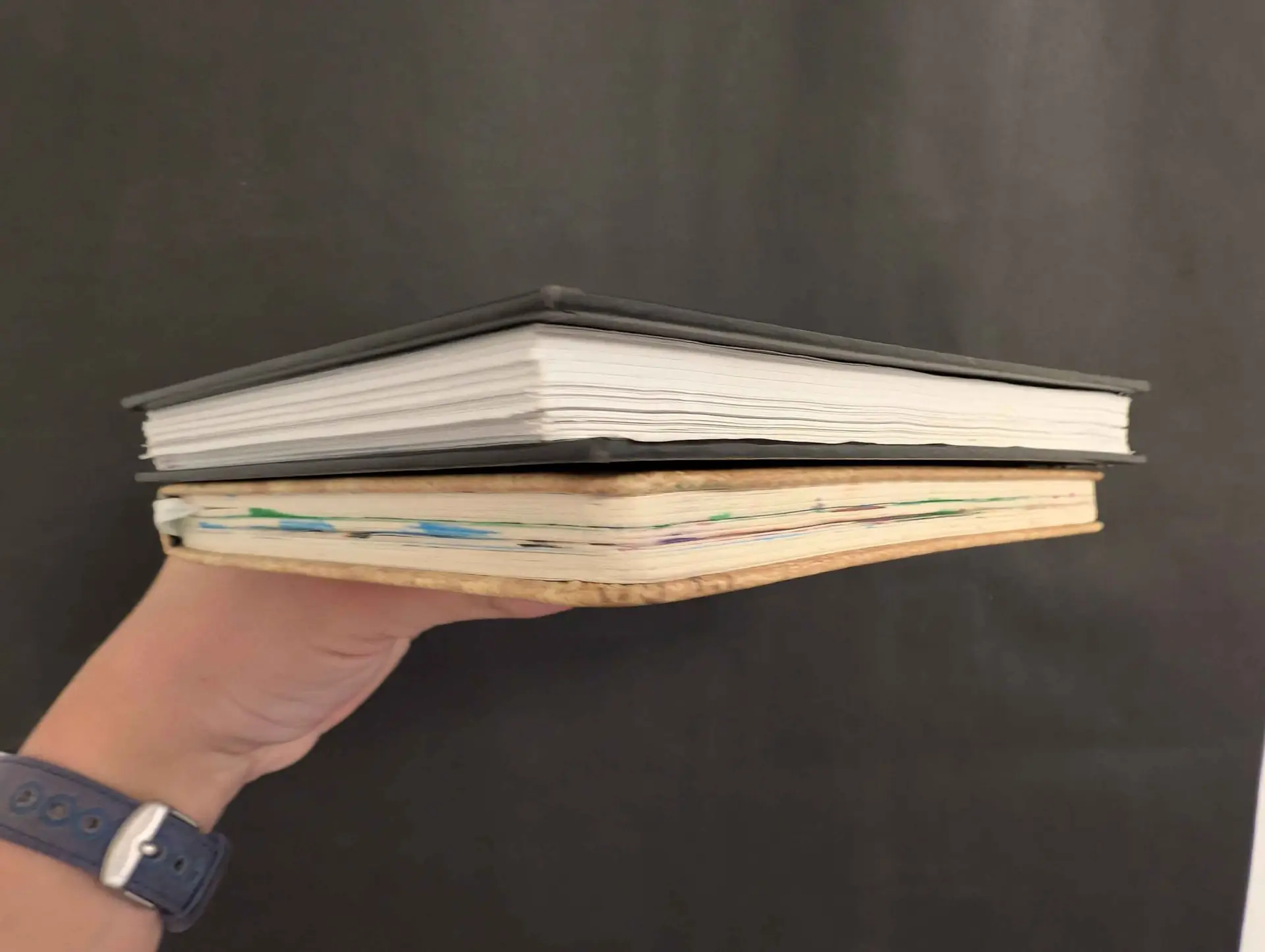 A man's hand holding up two art notebooks, one with clean page edges and the other colorful pages.